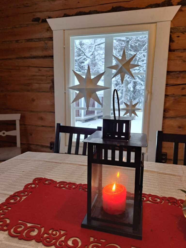 Pöydällä on punainen koristepöytäliina ja sen päällä palaa punainen kynttilä lyhdyssä. Ikkunassa roikkuu jouluisia paperitähtiä.
